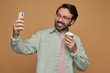 Studio portrait of bearded man standing over beige background wears casual shirt and glasses making...