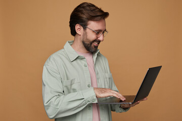 Studio portrait of bearded man standing over beige background wears casual shirt and glasses typing...