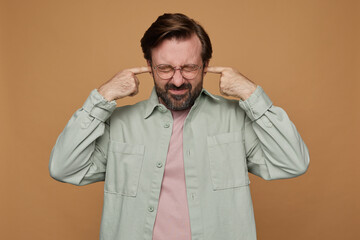Fototapeta premium Studio portrait of bearded man standing over beige background wears casual shirt and glasses grimaced, plug ears with a fingers with irritated facial expression