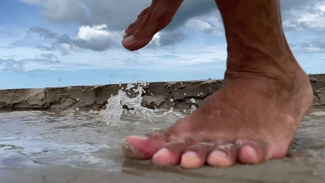 Man running barefoot on beach close up slow motion seafront splash of water with beautiful blue sky and cloud in background. Male feet step on sand - day