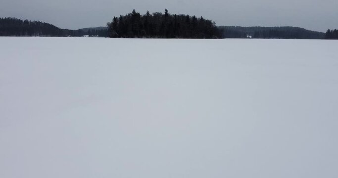 Flying over frozen lake and going up and over a little island in the frozen lake. Frozen lake with a island. Snow covered trees. Twilight time. Moody dark weather. Winter season. Drone footage. 