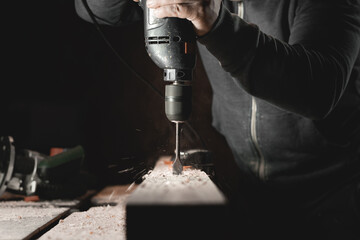A man works with a drill in his workshop. Carpenter drills with a hand power tool in a dark room...