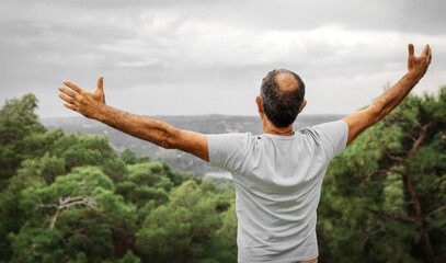 Fototapeta na wymiar An elderly 60-years old man standing with opened arms and looking on skyline on mountain and forest background