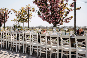 Long wooden table decorated with flowers and wooden chairs at an outdoors wedding party in a garden.