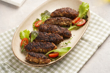 Several homemade minced meat sausages köfte on an oval beige colored plate with green leaves and cherry tomatoes on green checkered kitchen towel