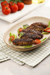 Several homemade roasted minced meat sausages köfte on an oval beige colored plate with green leaves and cherry tomatoes on green checkered kitchen towel