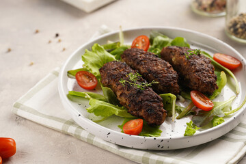 Several homemade roasted minced meat sausages köfte on a round white plate with green leaves and cherry tomatoes on beige colored linen napkin