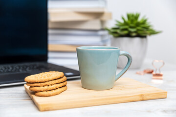 Blue cup and cereal cookies on a wooden board. Blurred computer and stack of books in the background