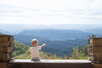 rear view blonde kid boy sitting outdoors on the bench and enjoying beautiful nature view