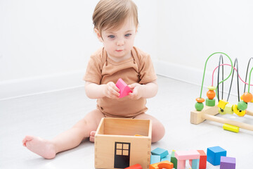 cute baby girl in brown bodysuit playing with colorful wooden toys on white room