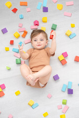 cute smiling baby girl in brown bodysuit laying on floor around colorful wooden blocks. top view