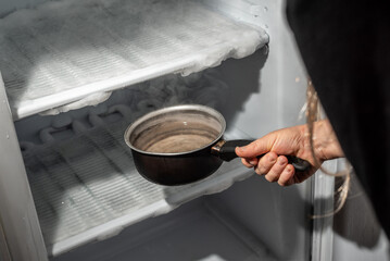 Hand introducing a saucepan with hot water to defrost the ice in the fridge