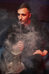 handsome man smokes a hookah in a cloud of white smoke, holds a mouthpiece in his hands