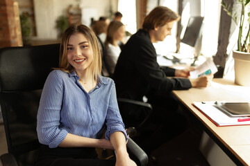 Attractive young confident businesswoman sitting at office desk with group of colleagues in background. Concept of success, business, career, achievements