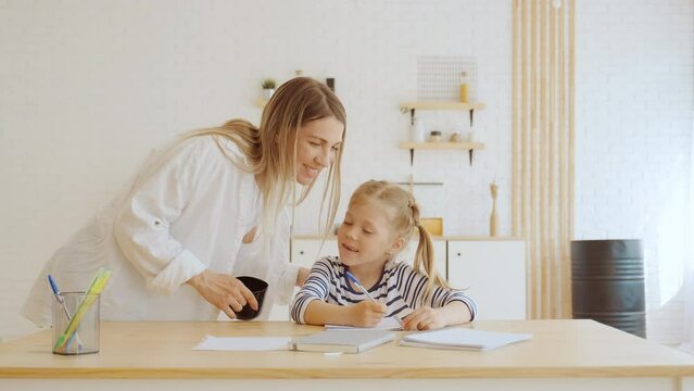 Little preschool girl doing homework studying alphabet, writing in notebook. Her mother helps her and supports, checking her school task, then kisses daughter and hugs her. Living room at home