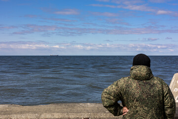 A man in soft focus in khaki clothing stands with his back and looks at the cold sea. A ship sails in the distance. A clear sunny day with a blue sky with clouds in Baltiysk, Kalinigrad region, Russia