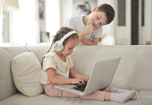 little girl works with a computer sitting on the sofa while her brother watches
