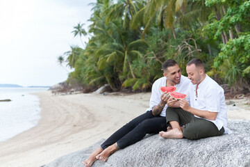Gay couple sitting on a tropical beach while eating watermelon