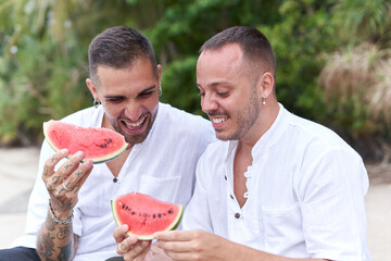 Two gay men sitting on a beach while smiling and holing a piece of watermelon