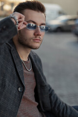 Street portrait of a handsome young hipster man in fashionable casual business clothes wearing stylish blue sunglasses and sitting in the city