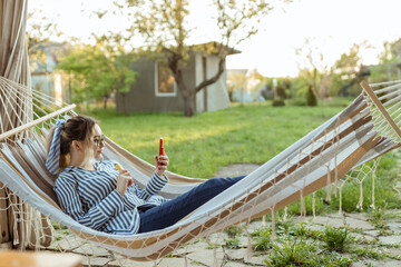 A beautiful girl in a hammock smokes an electrode cigarette and uses a smartphone outdoor