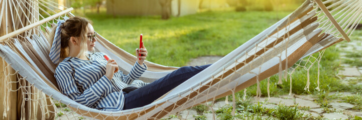 A beautiful girl in a hammock smokes an electrode cigarette and uses a smartphone outdoor
