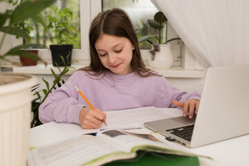 the girl studies online in front of a laptop, virtual distance learning, does homework online, studies at home in class, sitting at a table with a laptop.