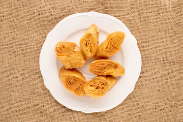 A few pieces of sweet honey baklava with a white ceramic plate on burlap, close-up, top view.