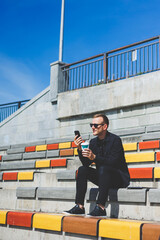 Photo of smiling businessman in suit holding mobile phone while standing outdoor near building with takeaway coffee