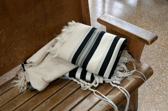 A tallit Jewish prayer shawl with traditional knotted strings on the four corners sits on a bench in a synagogue.