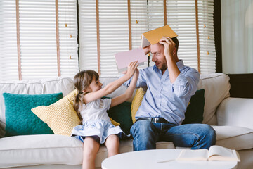 Happy father and cute daughter sitting on sofa and holding book on heads and looking each other....