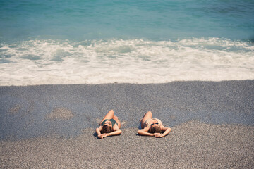 Fototapeta na wymiar Two girls girlfriend are sitting on the sea sandy shore and the waves soaked them in bathing suits on a sunny warm day