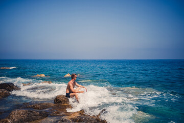 A young man stands on the rocks overlooking the open Mediterranean Sea. A guy on a warm summer sunny day looks at the sea breeze
