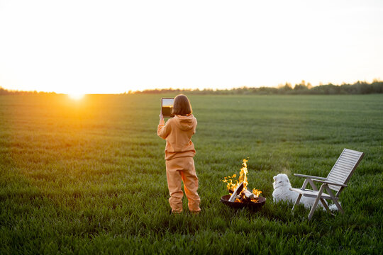 Young person takes photo of beautiful sunset while relaxing with pet by the fireplace on green field. Spending summer time and enjoying nature concept