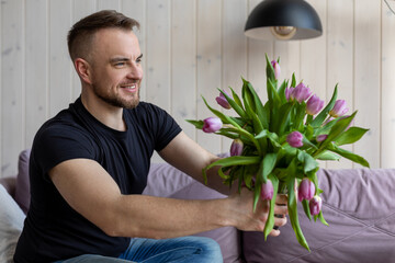Gute smiling man in black T-shirt and jeans is sitting at home on sofa with bouquet of tulips and preparing surprise for his girlfriend. Preparation for date. Attention and care.