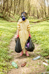 Woman in gas mask and protective clothes stands with garbage bags collecting scattered plastic in the woods. Problem of environmental pollution. Safe the world concept