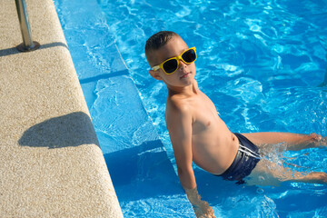 A little boy is swimming in the outdoor pool. The child is floats in an sunglasses.
