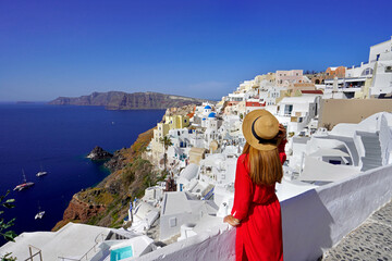 Beautiful woman in Oia village holds hat when looking cityscape from terrace in Santorini Island,...