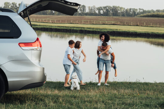 Happy Four Members Family Having Fun Time Outdoors By The River, Mother And Father With Two Teenage Children And Pet Dog Enjoying Road Trip On Minivan Car