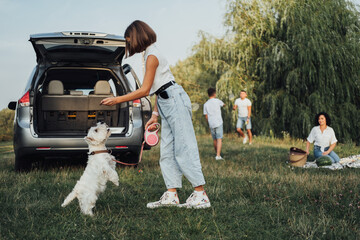 Teenage Girl Playing with West Highland White Terrier Dog on Background of Her Family Having Fun Time Outdoors on a Road Trip by Minivan Car