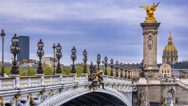 The ornate Pont Alexandre III in the downtown of Paris, France with the golden dome of the Invalides and the Tour Montparnasse in the background below an overcast sky