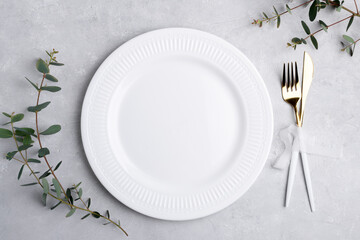 Festive table setting with white classic plate, golden cutlery set and fresh eucalyptus leaves on grey concrete table top view. Plate mockup. Copy space, element for design