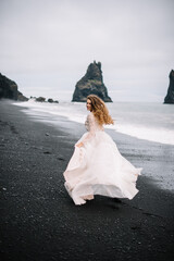 Bride in old classy wedding dress are running on the Black Sand beach in Vic, Iceland with rock islands on background.
