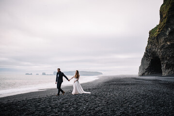 Bride and groom in wedding outfits walk on the Black Sand beach with the view to the Atlantic ocean and horizon. Epic view in Iceland Elopement.
