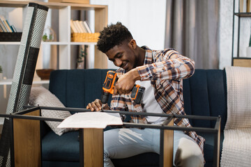 African american man sitting on cozy sofa and using cordless drill for assembling shelf at living...
