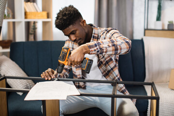 Portrait of happy african american man in casual attire with cordless screwdriver in his hand assembling modern cupboard at living room. Moving in new house concept. - obrazy, fototapety, plakaty