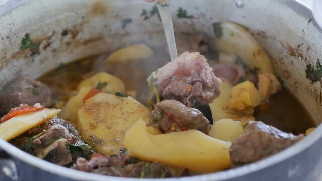 With a metal spoon, put pieces of hot meat and potatoes into a white bowl from which steam comes. Armenian khashlama. Close up, slow motion. National Armenian and Georgian food