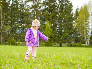 a child in a hat and jacket walks in the park on the grass against the background of trees