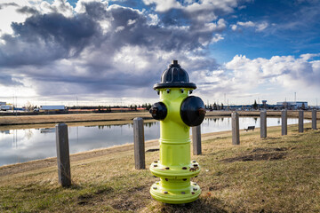 A yellow fire hydrant connected to infrastructure standing next to a storm retention pond in an industrial park in Airdrie Alberta Canada under a dramatic sky. - Powered by Adobe