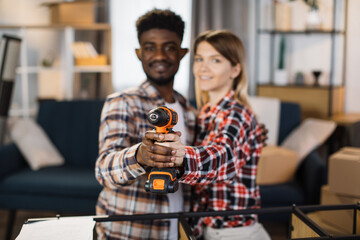 Beautiful and happy multiracial family posing at their new apartment with cordless electric drill in hands. Focus on repair tool. Blur background.